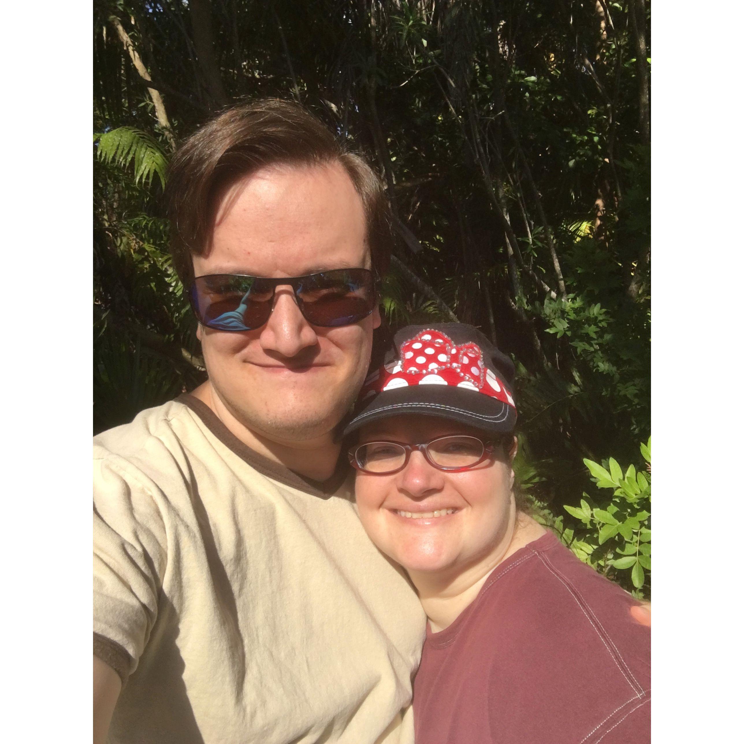 The couple smiling outside. Olivia wears a Minnie Mouse hat and Zac wears sunglasses.