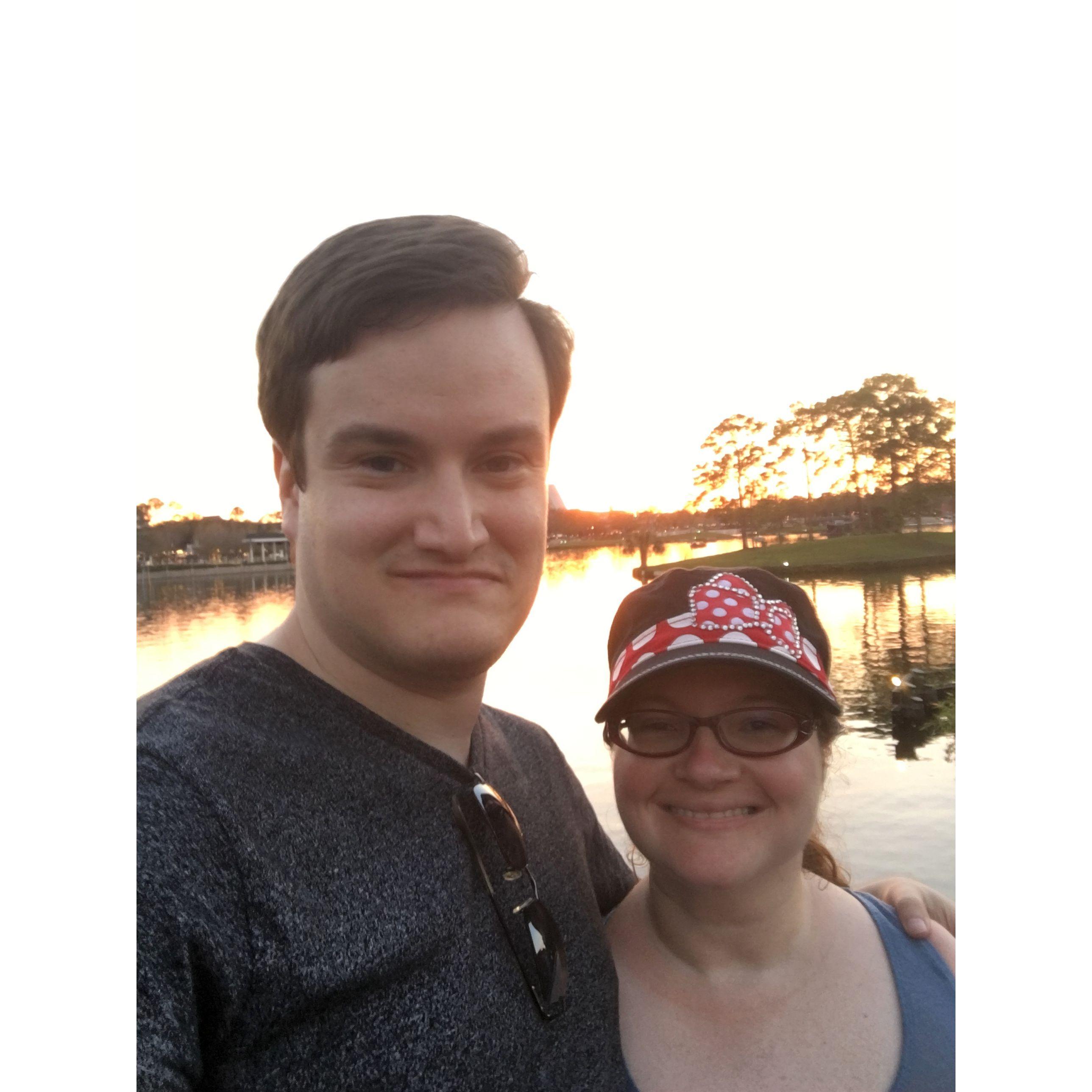 The couple smiling in front of a lake at Disney. Olivia wears a Minnie Mouse hat.