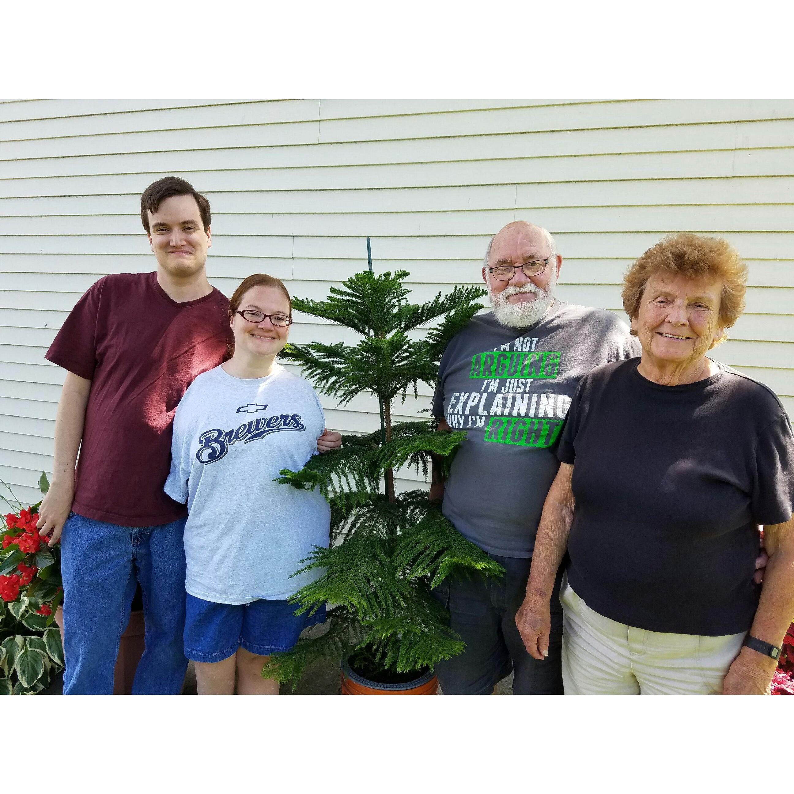 Zac, Olivia, and Olivia’s grandparents smiling next to an evergreen tree in her grandmother’s garden.
