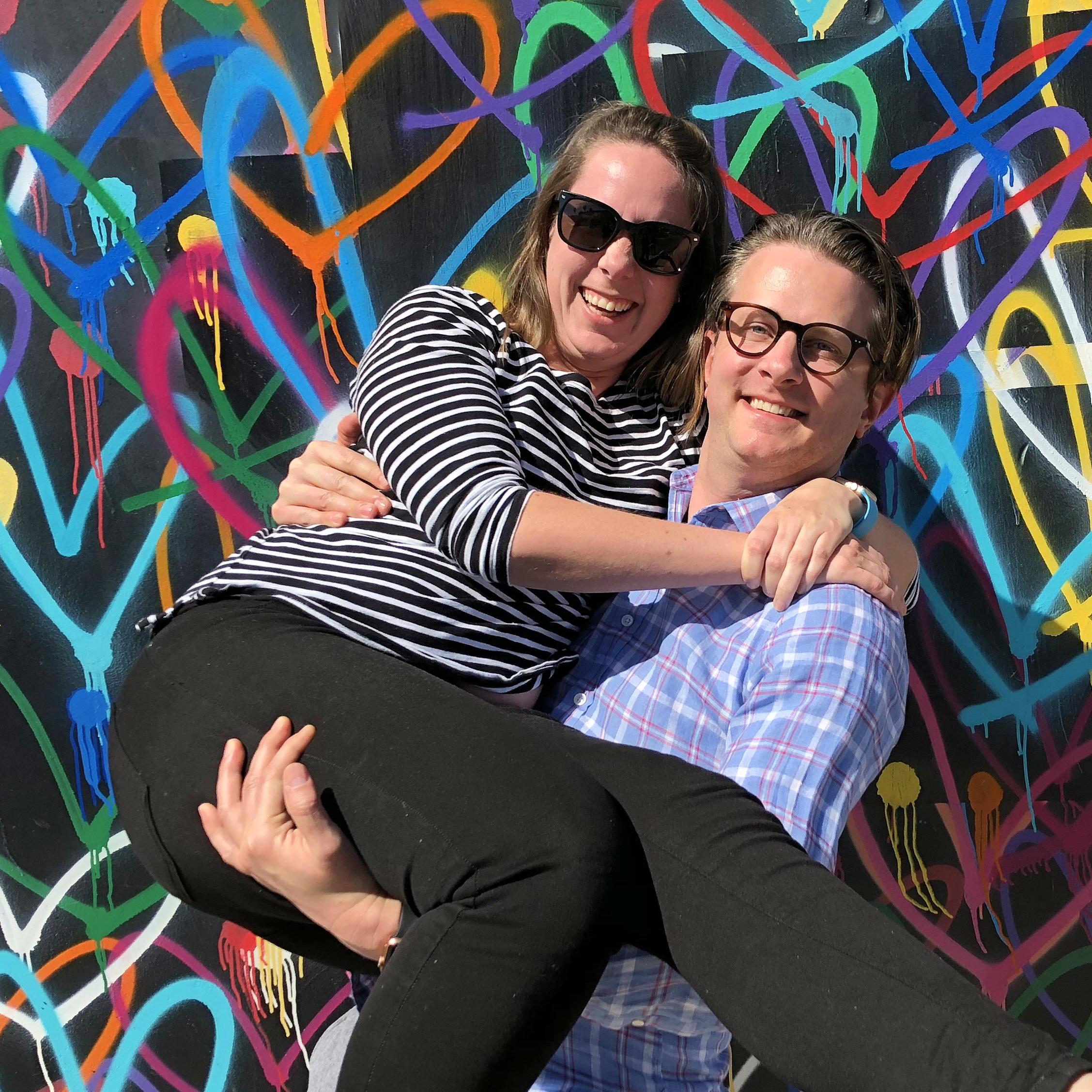 James carries Ruth for a picture in front of a wall of spray painted hearts.