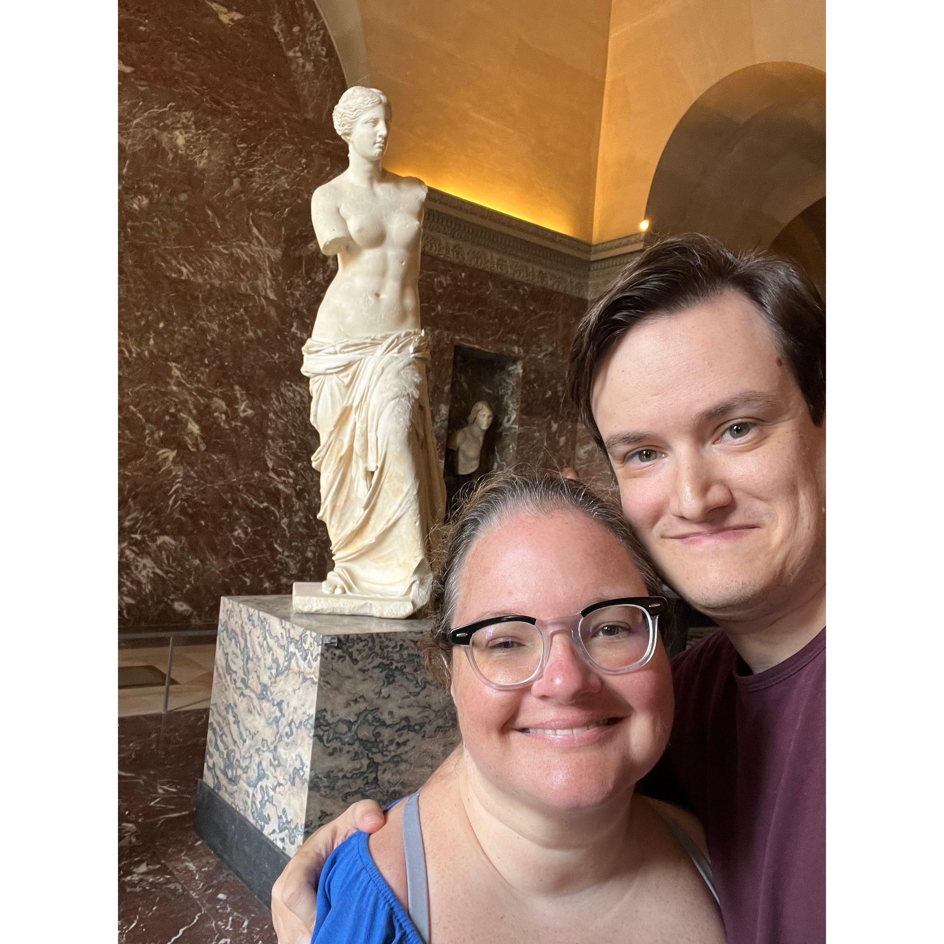 Selfie with the famous statue Venus de Milo behind them. The statue is of a woman with no arms.