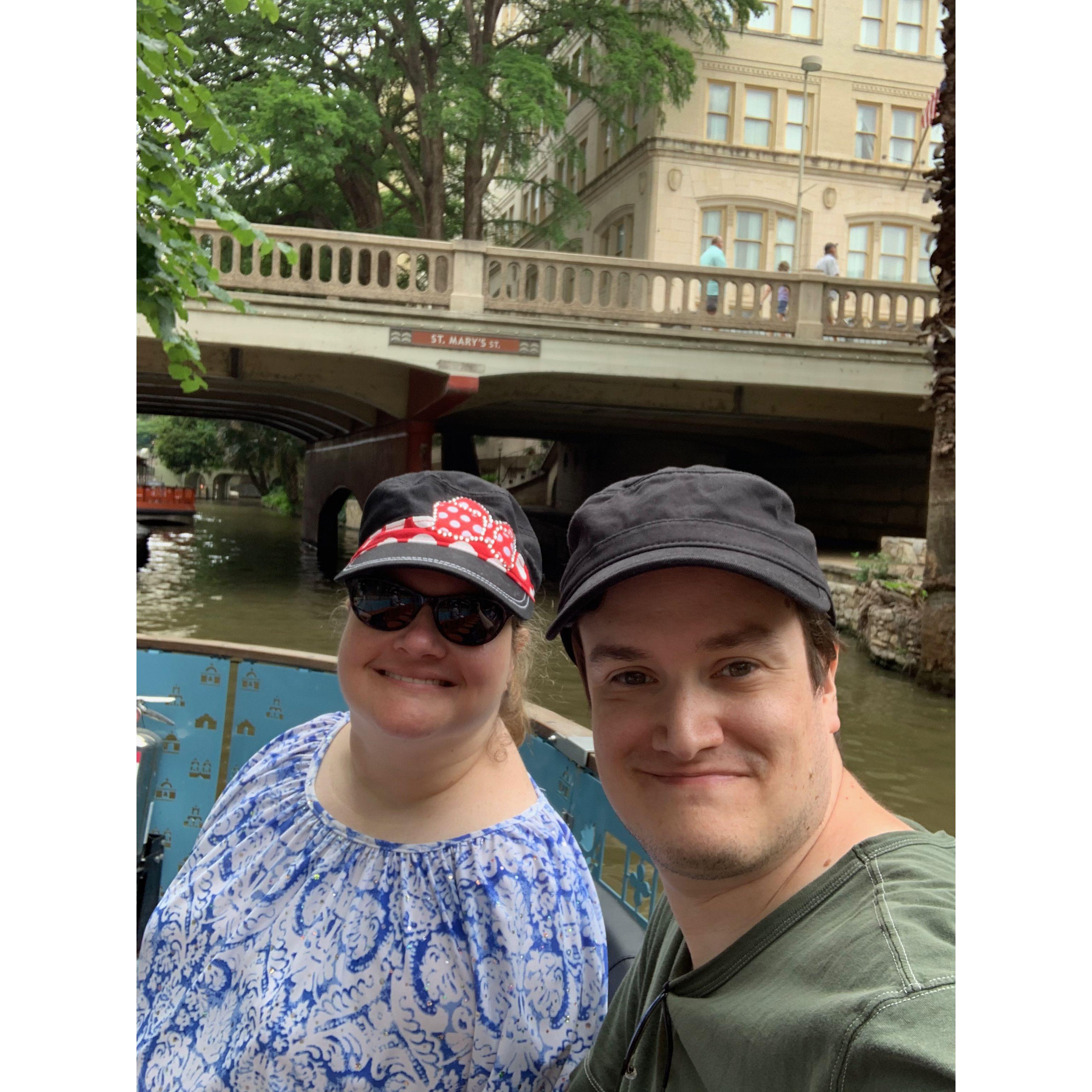 Olivia and Zac on a boat in San Antonio. They pass underneath a bridge that says “St. Mary’s street” surrounded by trees and old buildings.