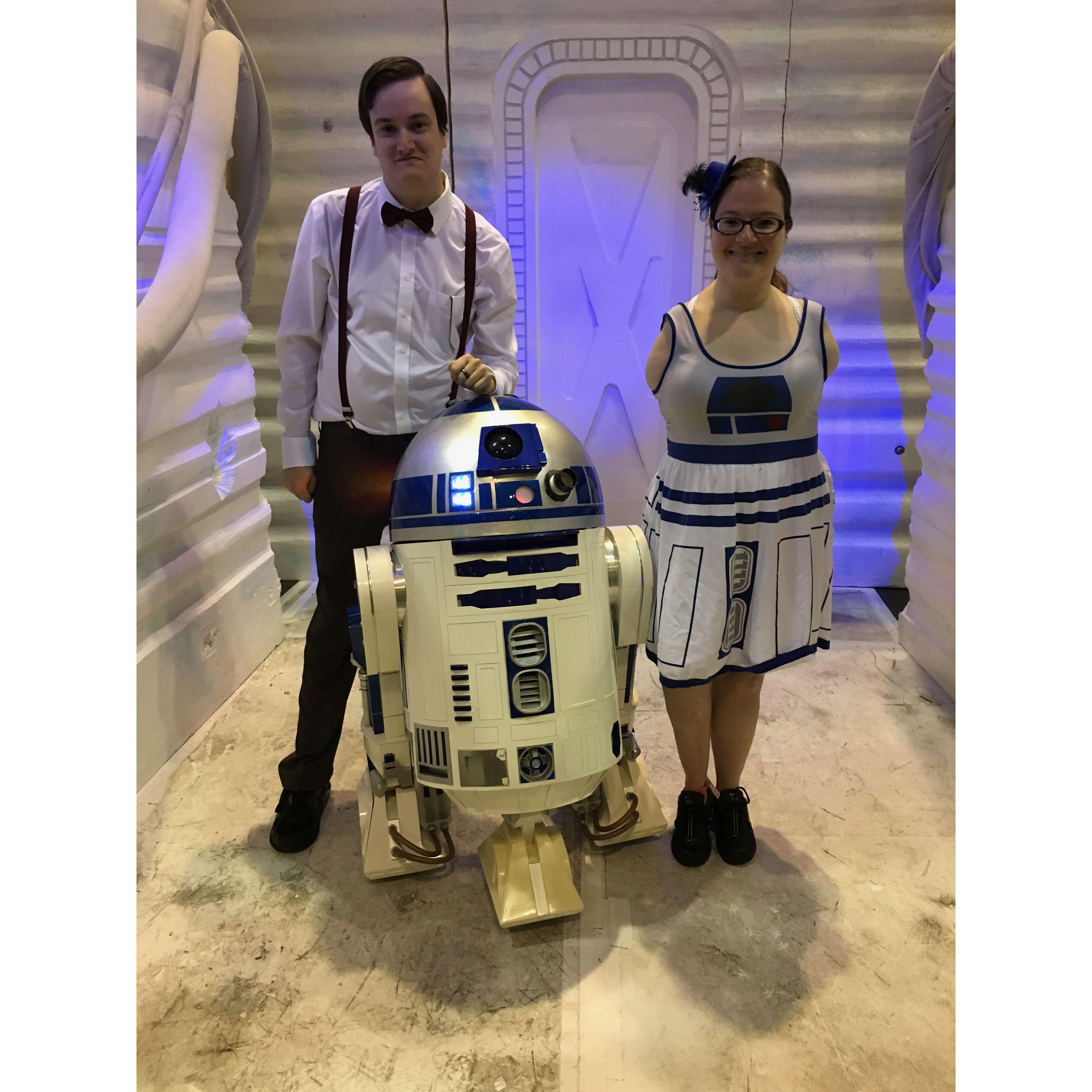Olivia and Zac posing next to Star Wars R2D2. Zac wears a white button up with a bow tie and suspenders. Olivia wears a dress that is designed to look like R2D2.