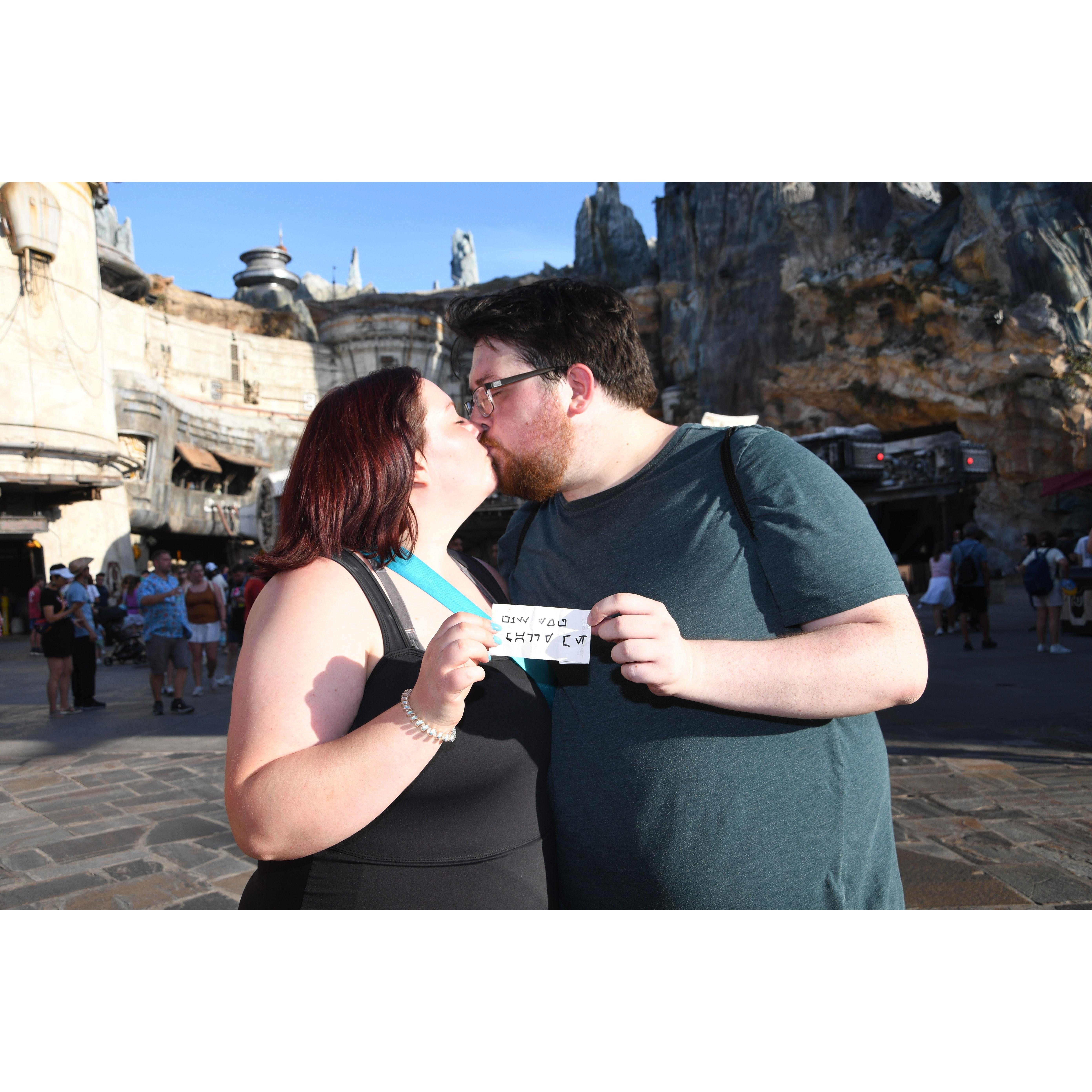 Sarah proposed on June 1st, 2024! Pictured here is the couple smooching over "Will you marry me?" translated into Aurabesh, the language of Batuu (AKA Disneyworld's Star-Wars-themed land).