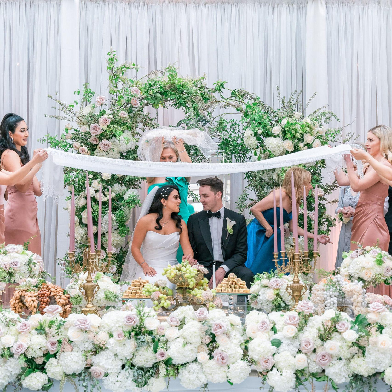 The couple sit under the white cloth and by their sofre-aghd spread and have sugar cones rubbed over their heads.
