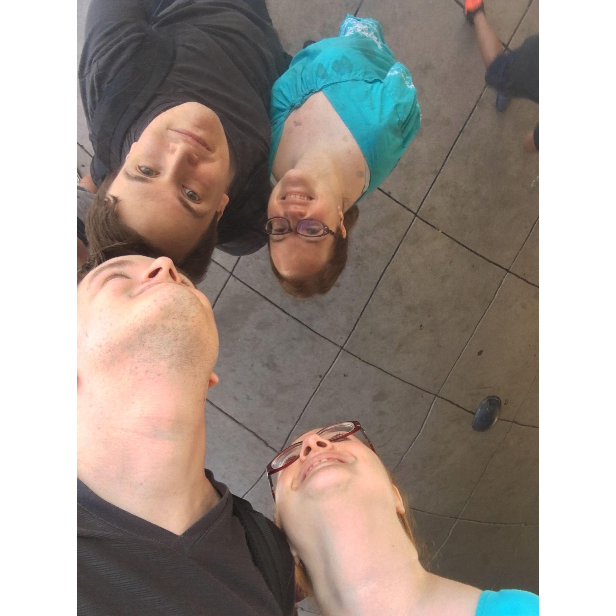 The couple smiling up in the bottom of the Chicago Bean. They take a selfie in the reflection