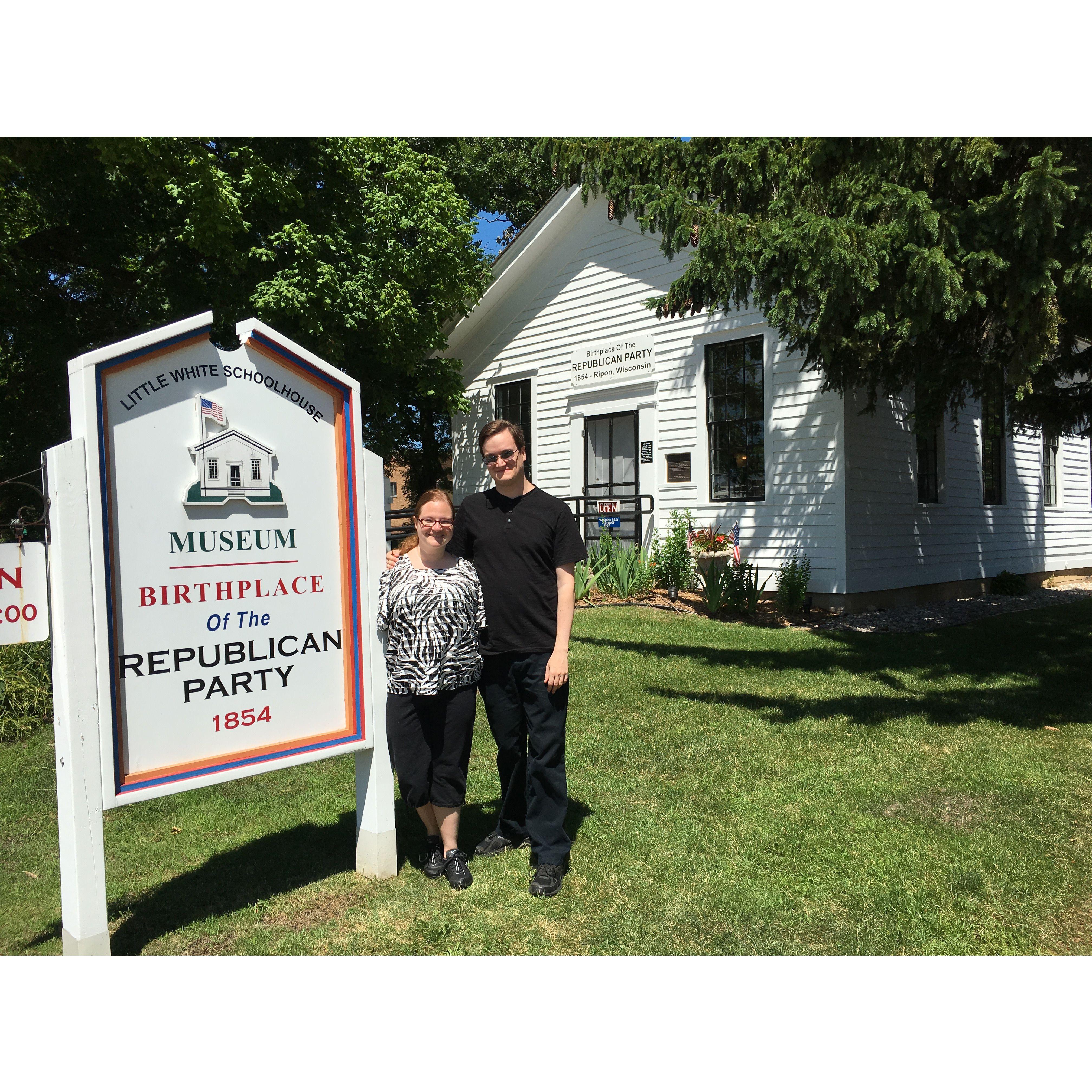 The couple posing next to a sign and building in Ripon,WI. The sign reads: little white school house museum. Birthplace of the Republican Party in 1854.