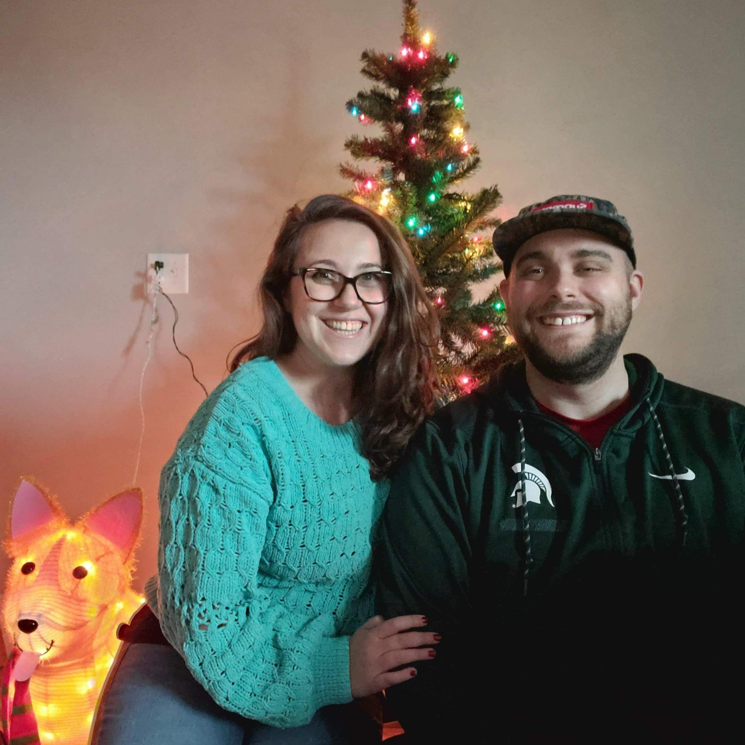 Our first Christmas photo together (Dec 2020)
Alt Text: Maeghan and Keiran sitting together in front of a small, artificial Christmas tree with our light up tinsel dog, Omelette!