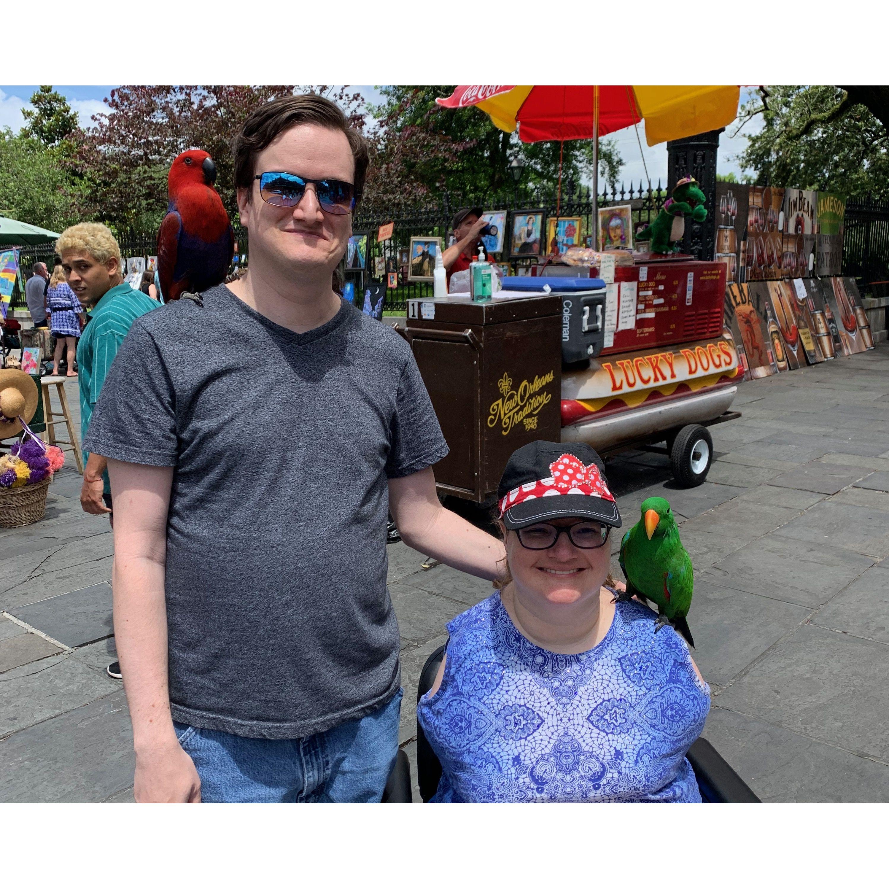 The couple smiling outdoors at an art fair in New Orleans. Olivia is seated in her wheelchair with a large green bird perched on her shoulder.