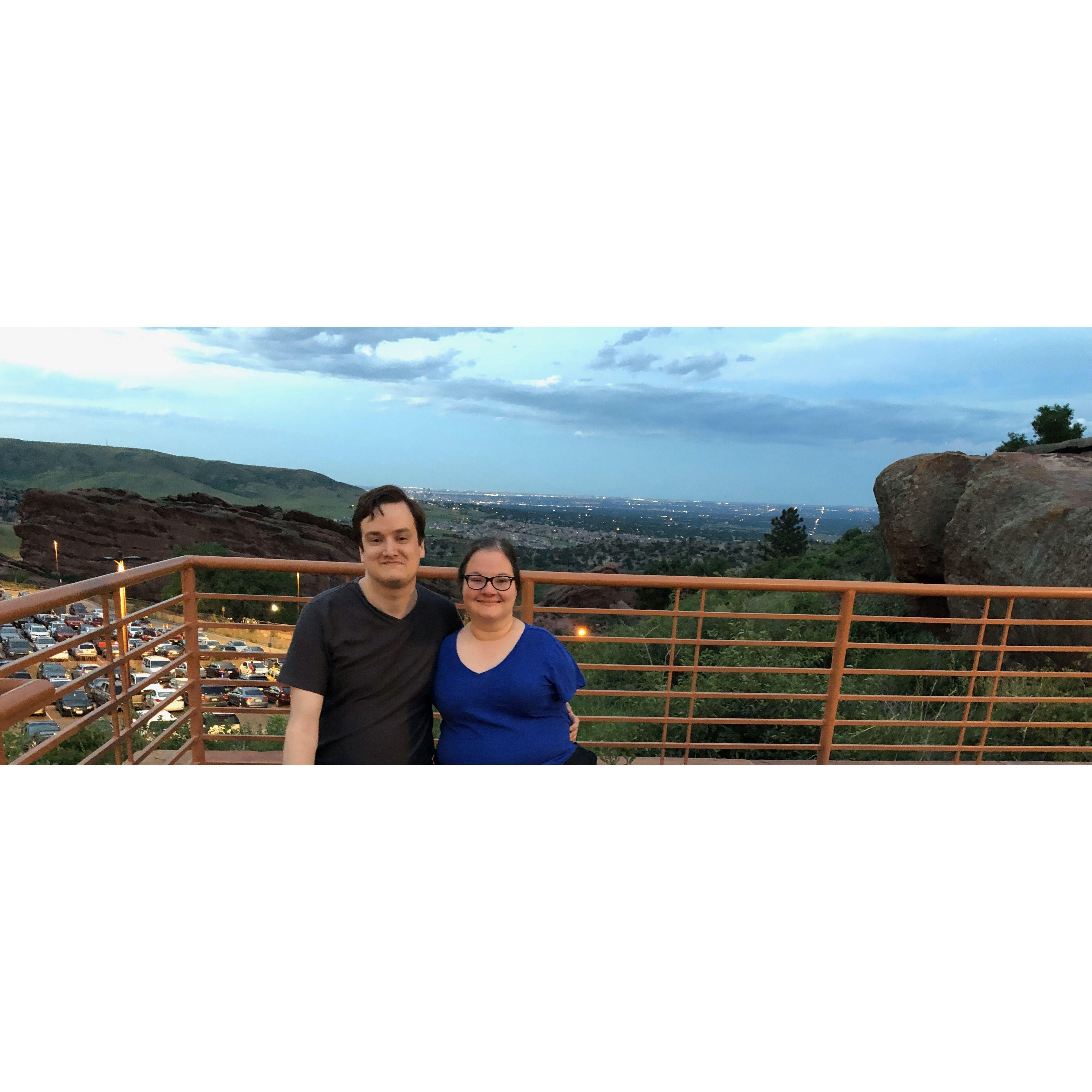 The couple standing at a scenic viewpoint at Red Rock Canyon with rolling hills, large rock formations and a setting sky behind them.
