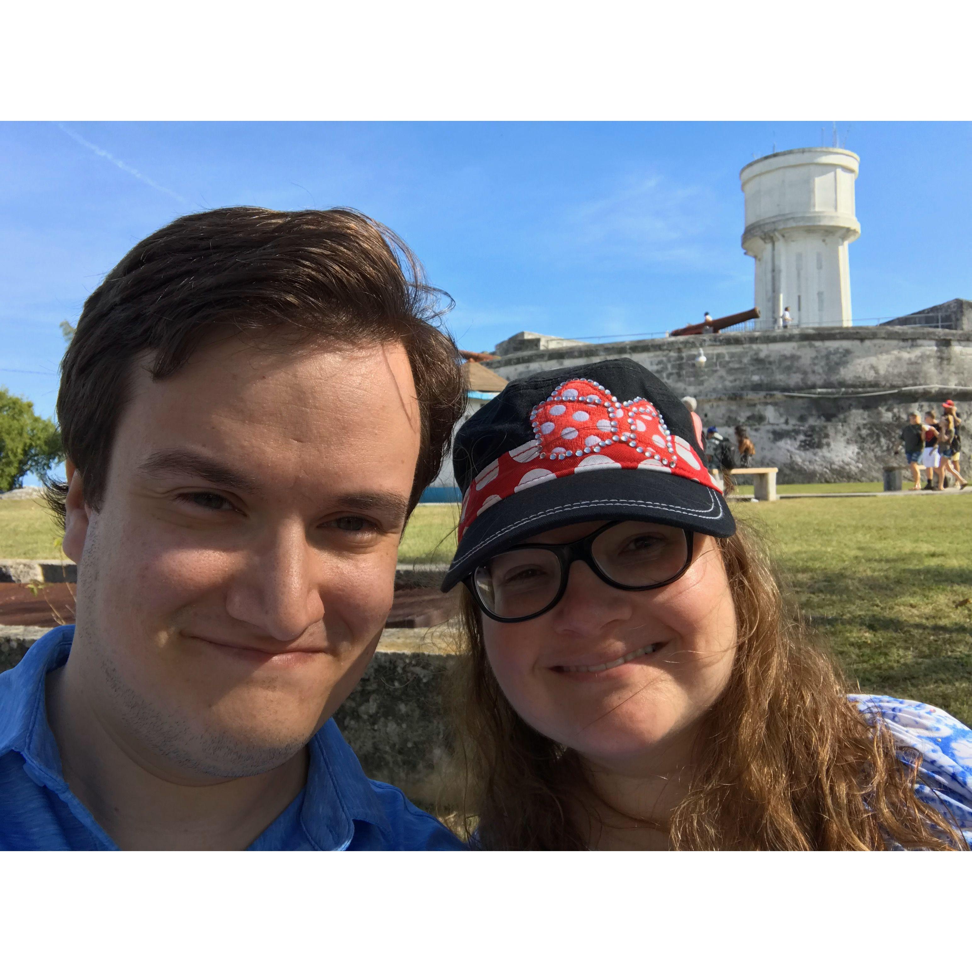 The couple smiling outside of an old fort in Nassau.. There is a tall wall, a cannon, and viewing post behind them.