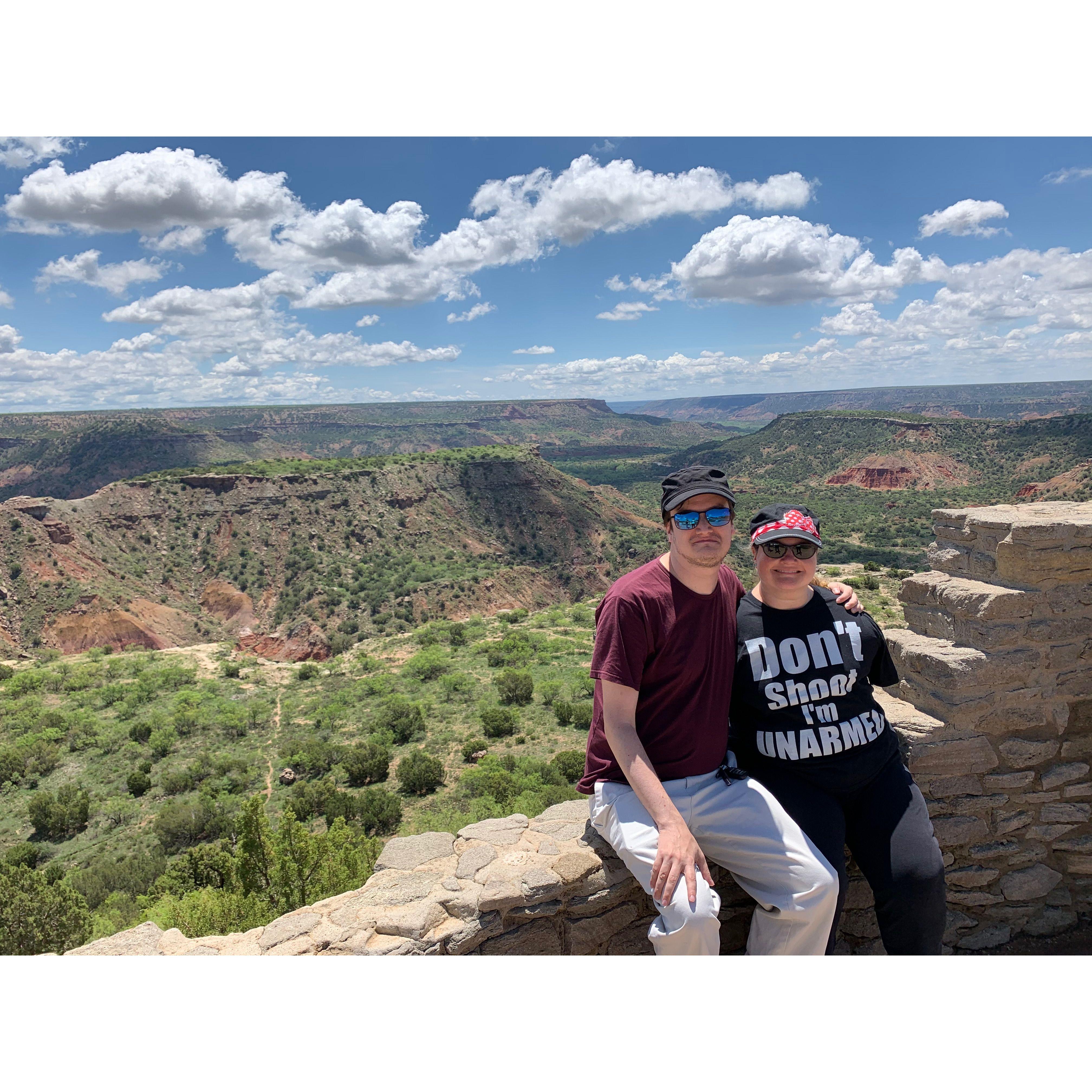 Couple seated on a rocky wall overlooking the Palo Duro canyon, with fluffy clouds in the sky. Olivia wears a funny shirt that reads “Don’t shoot I'm unarmed”.