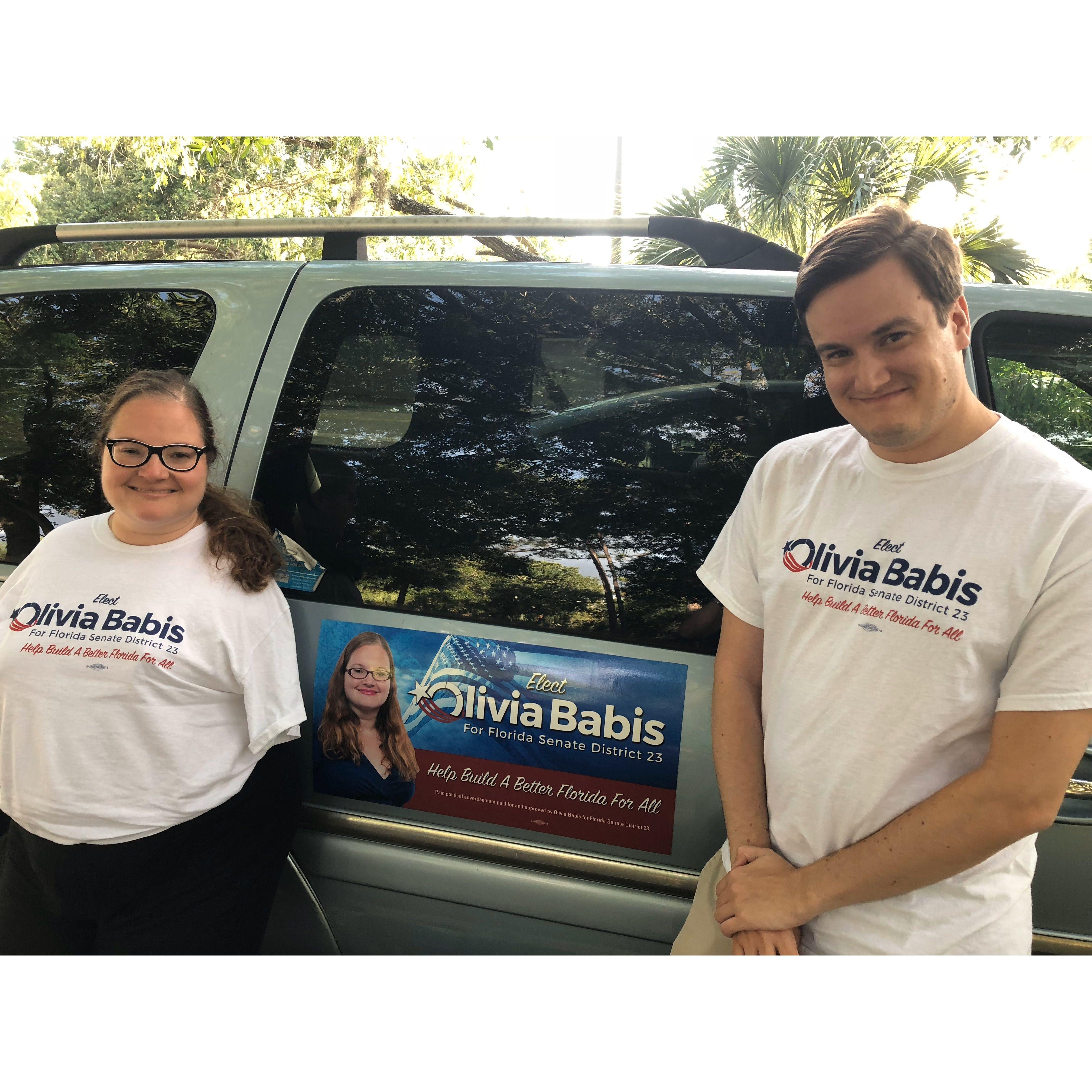 Olivia and Zac smiling in front of her campaign car. They wear Olivia themed shirts. Quote on shirts and car say: Elect Olivia Babis for Florida Senate District 23. Help build a better Florida for all