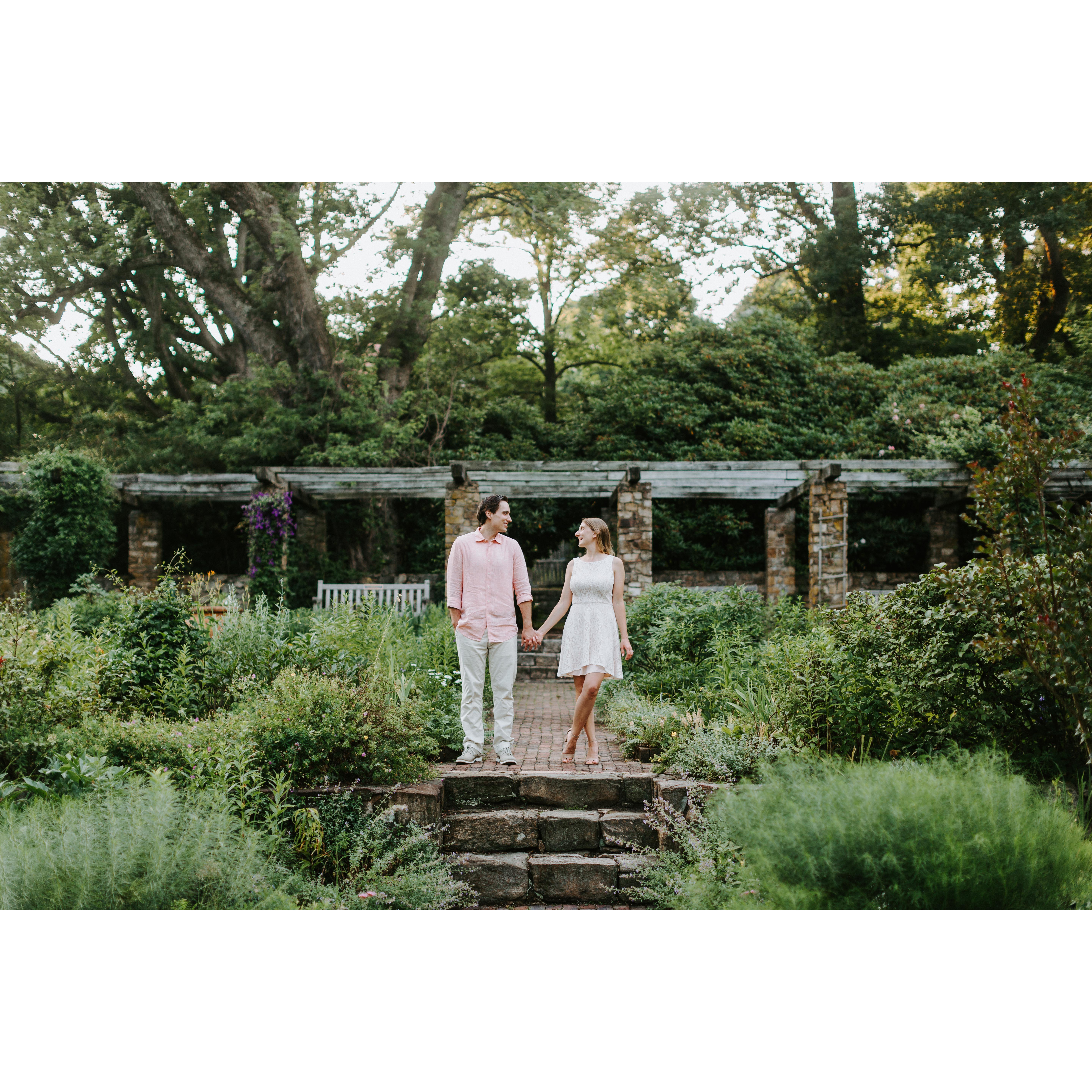 Matt and I holding hands. My one leg is crossed in front of the other. Matt's other hand is in his pocket. We are looking at each other.  Surrounded by a green garden with steps and a pavilion
