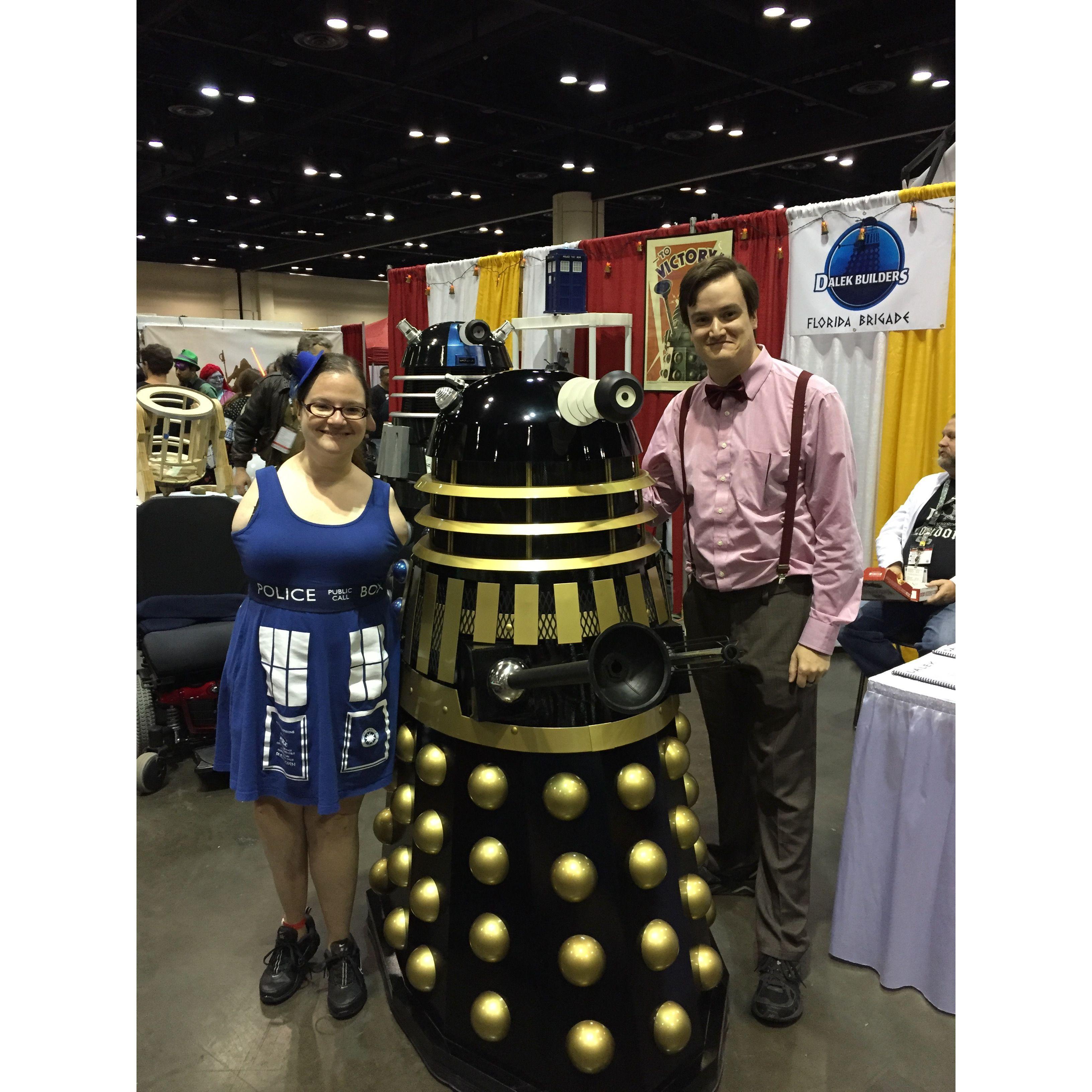 Olivia and Zac at a comic con event. They dress in Doctor Who fan outfits, with Olivia wearing a blue Tardis dress and hat designed to look like the police telephone booth. Zac is dressed as The Doctor. They pose next to a Dalek.