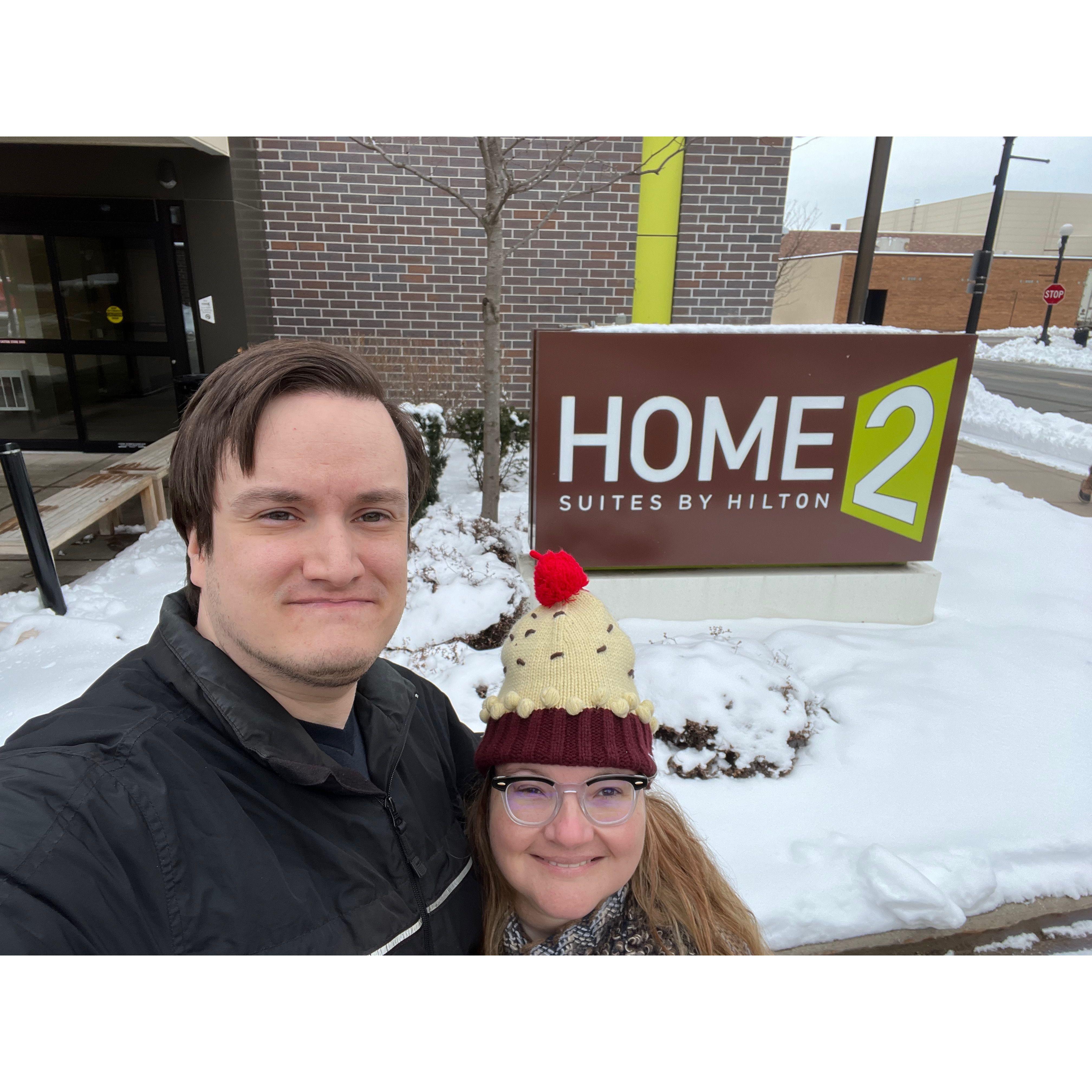 Selfie in front of Home 2 suites by hilton. There is snow on the ground and the couple is bundled up.