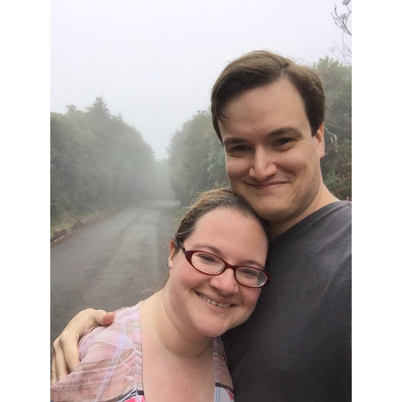 The couple outdoors on a path at Poas Volcano National Park. The air is foggy. Olivia leans her head on Zac’s chest and they both smile