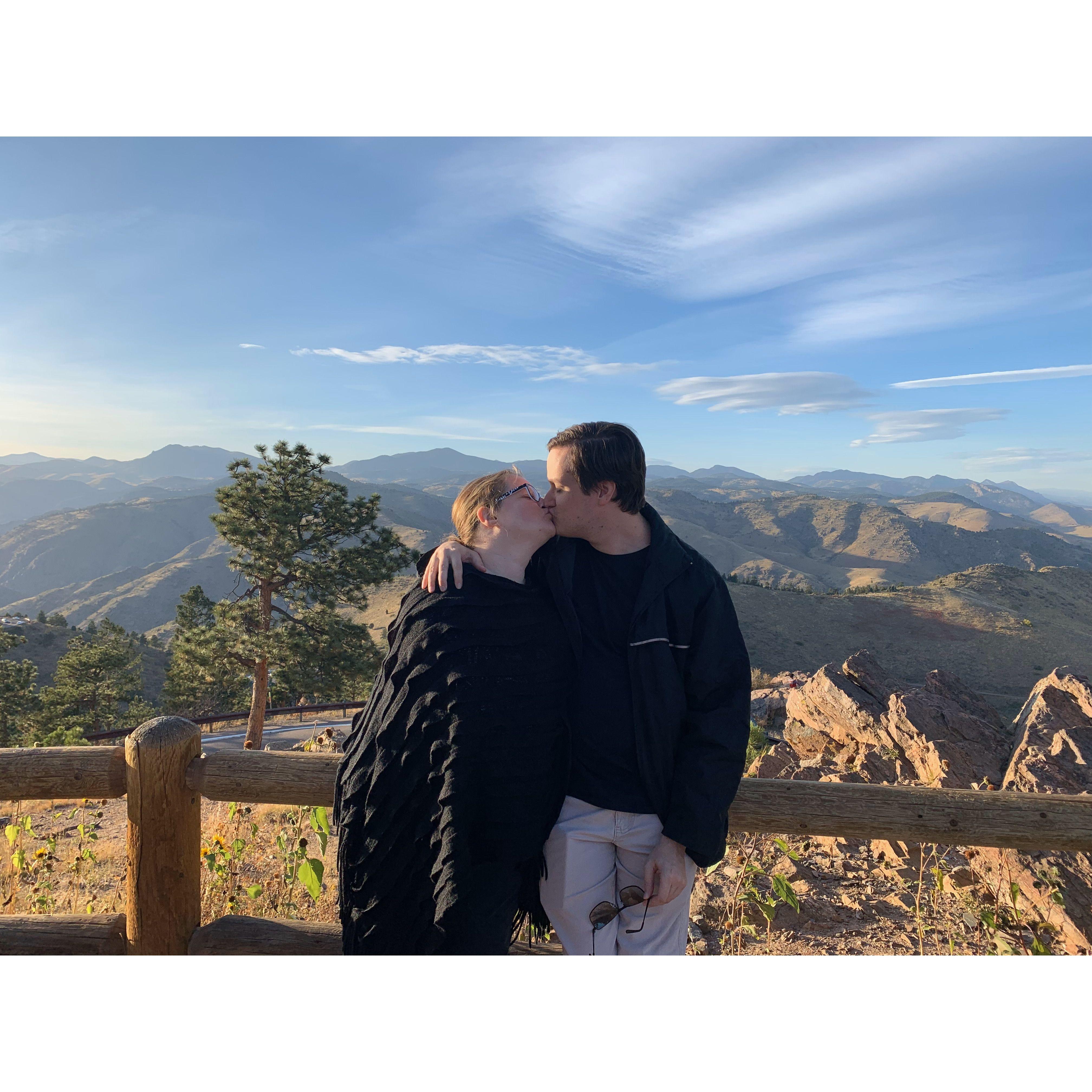 The couple standing at a scenic viewpoint at Lookout Mountain Park, CO, kissing. Behind them are arid trees and plants with mountains.