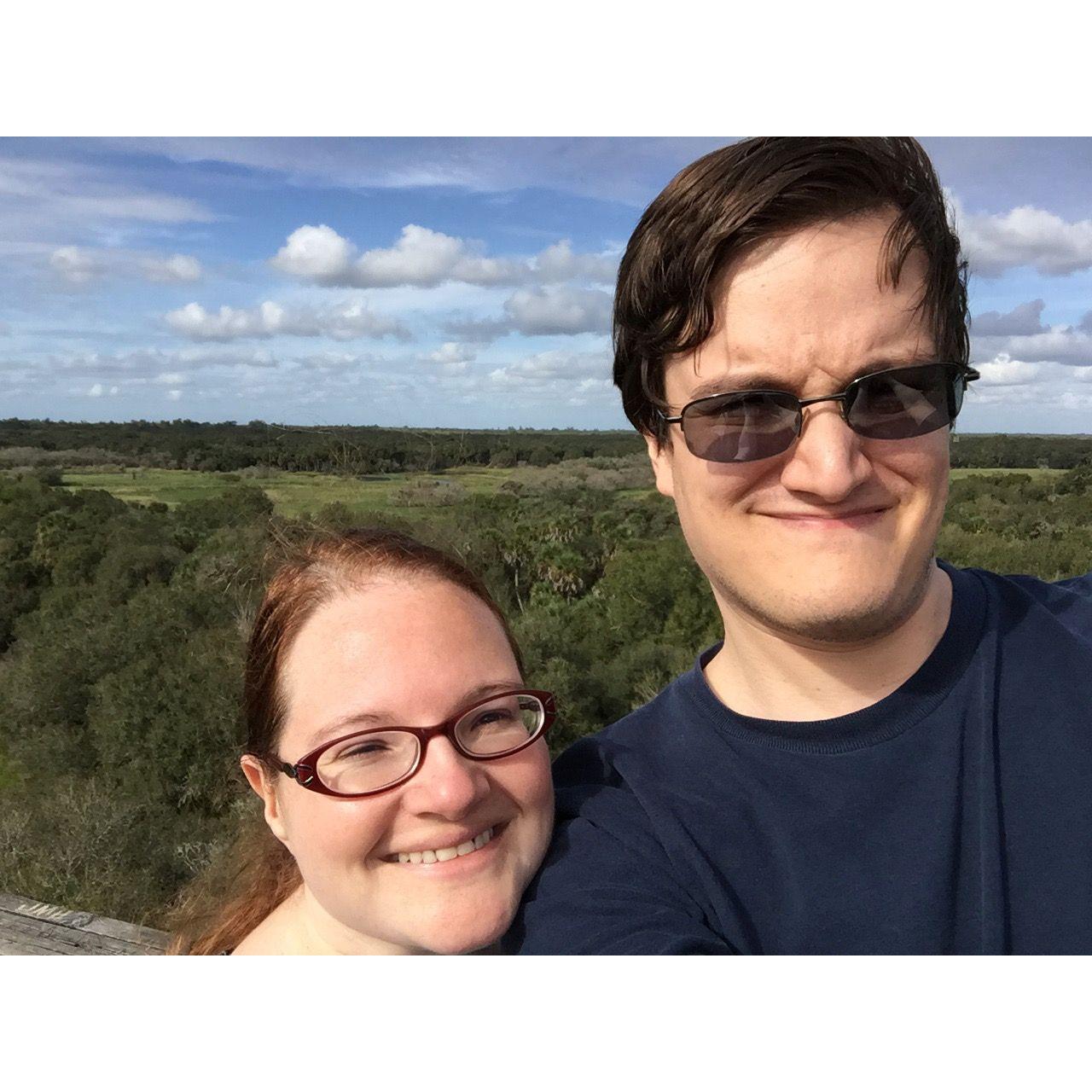 The couple take a selfie at the top of the Canopy Walkway at Myakka State Park. The background is picturesque with lots of trees and fluffy clouds